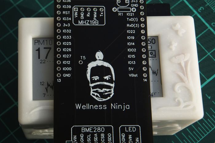 A custom circuit board silkscreened with a ninja face and the words Wellness Ninja, resting against two white resin enclosures.
