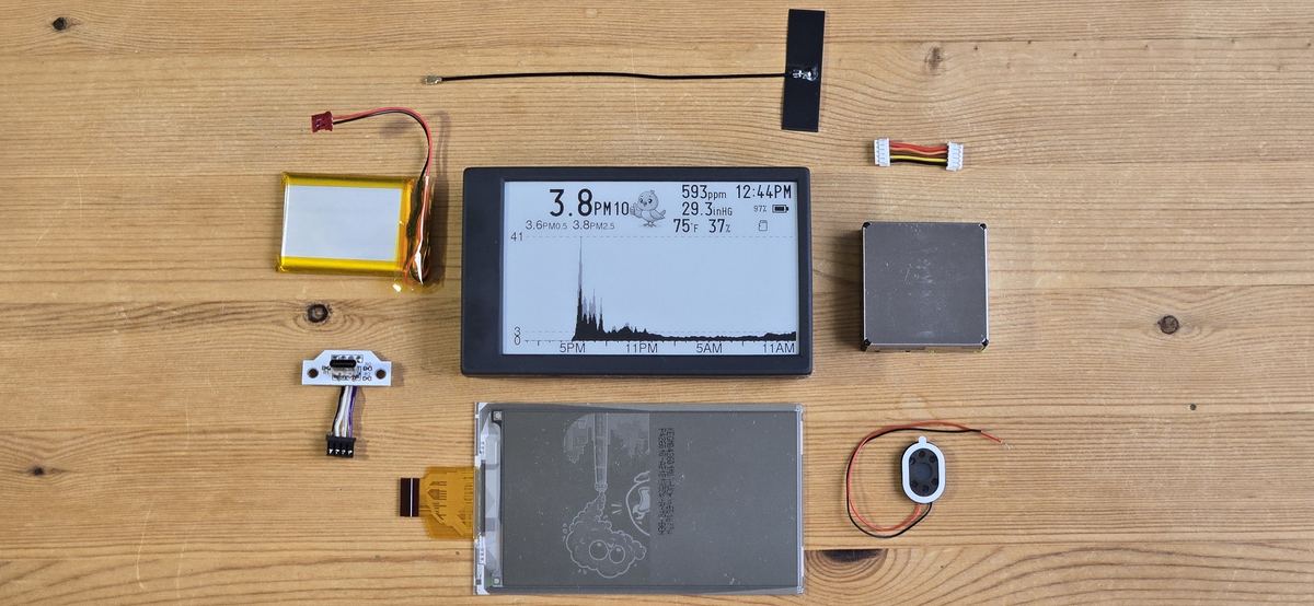 A Canary laid out in parts on a wooden bench: the assembled unit, a spare e-paper panel, battery, antenna, USB-C board, sensor module, speaker and ribbon cables.